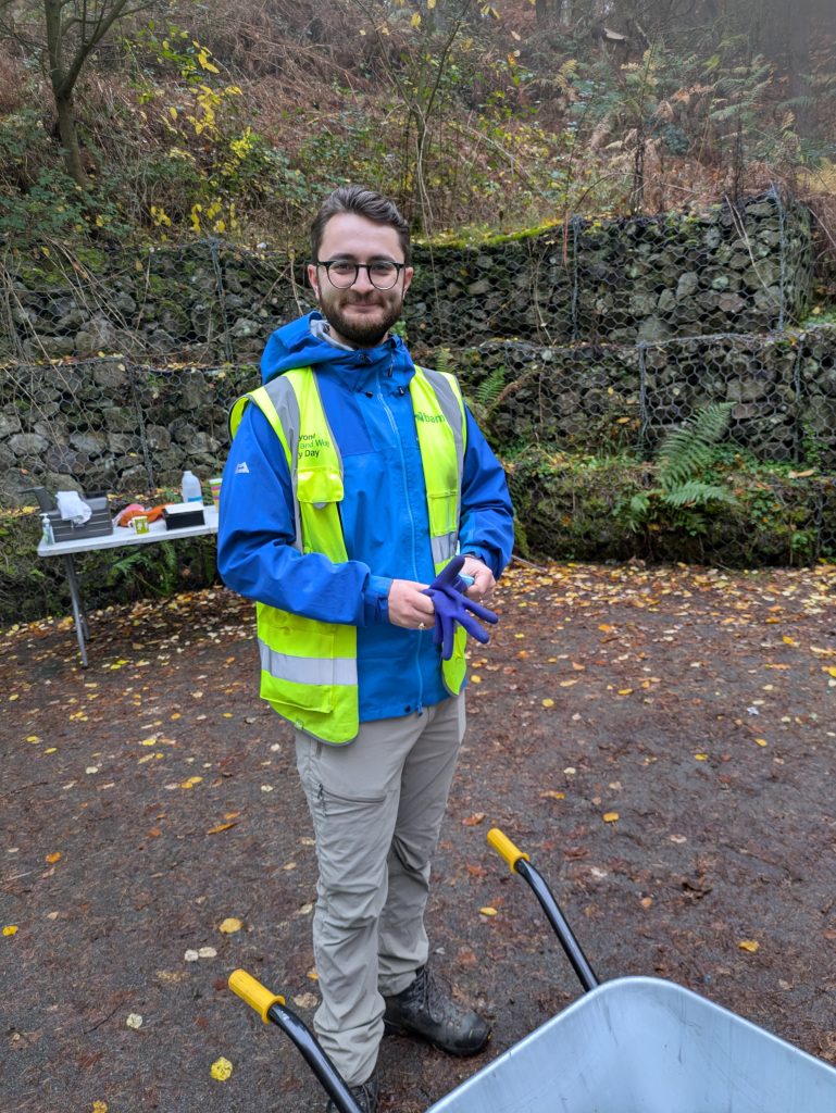 Tom Wimpenny, a bearded man in a blue jacket wearing hi viz standing next to a wheelbarrow in Huddersfield Riverside Nature Park